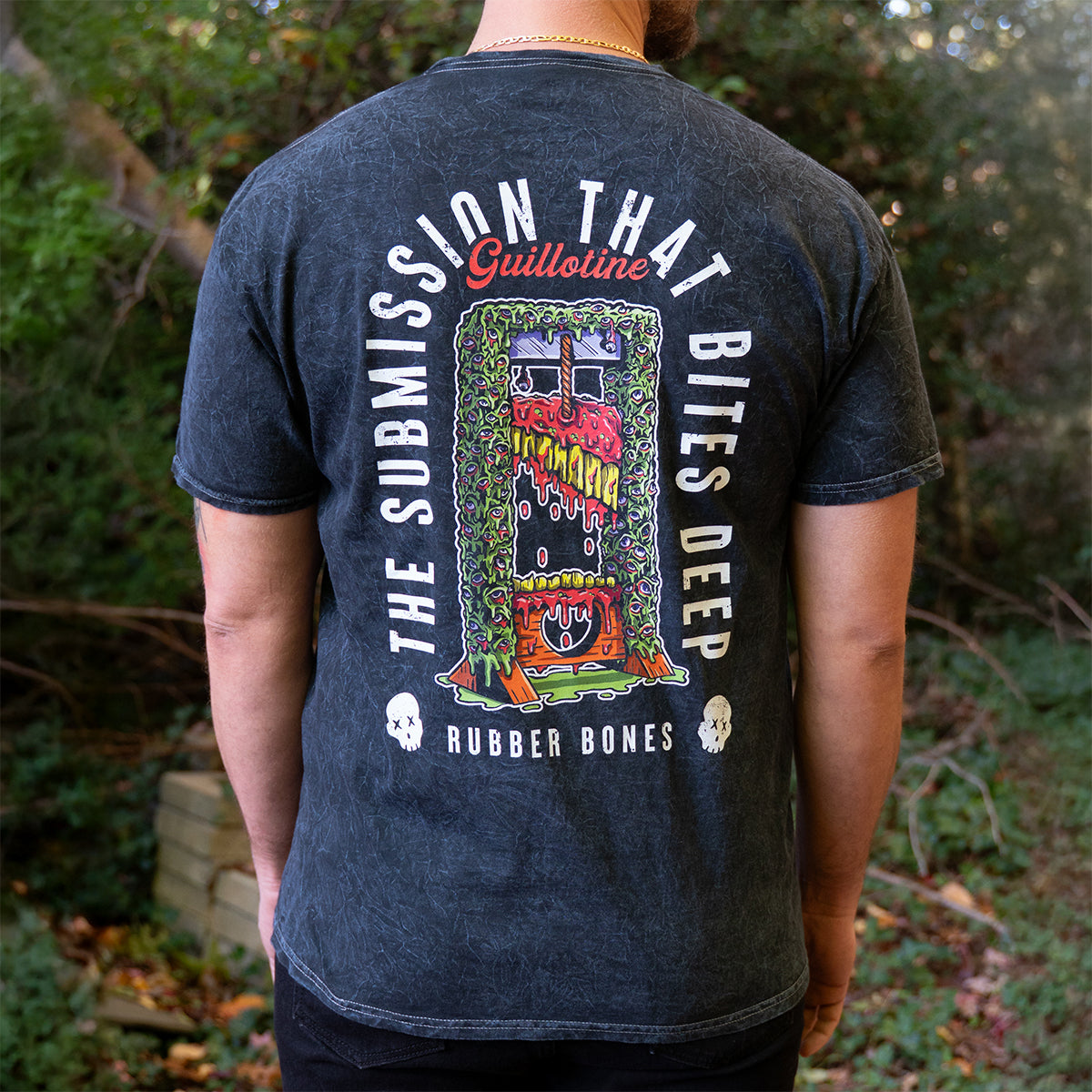 Back view of Rubber Bones Guillotine T-shirt featuring colorful guillotine artwork and text reading “The Submission That Bites Deep.”