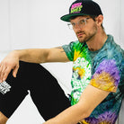 A male model sits against a white wall wearing a vibrant tie-dye “Legalize Leg Locks” t-shirt with neon green graphics, paired with black Rubber Bones shorts and a black snapback cap featuring the Rubber Bones logo in pink and green. He has a relaxed pose, clear-framed glasses, and a confident expression, embodying the rebellious and artistic vibe of Rubber Bones apparel.