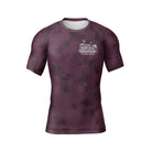 Maroon short-sleeve BJJ rash guard with a subtle black tie-dye pattern. A small white chest logo displays 'The Natural Grappler' and 'Rubber Bones' in a hand-drawn, artistic style.