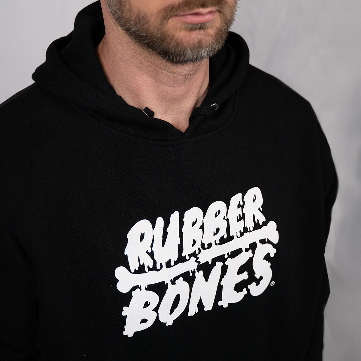 Man wearing black Rubber Bones logo hoodie showing white skull graphic and text on center chest, with double-drawstring hood visible.