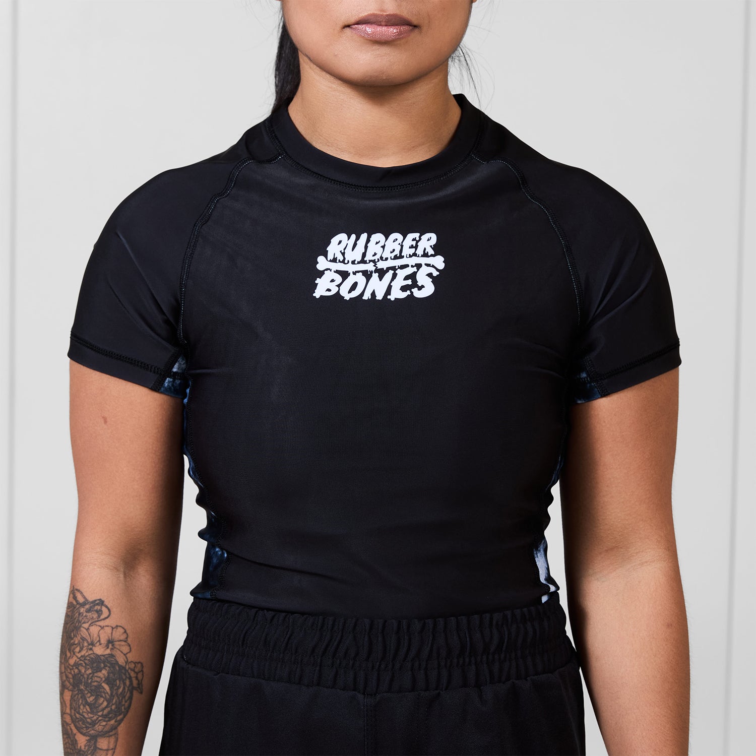 Model wearing the Women’s Ash BJJ Rash Guard featuring black body, white Rubber Bones logo, and gray marble side details.