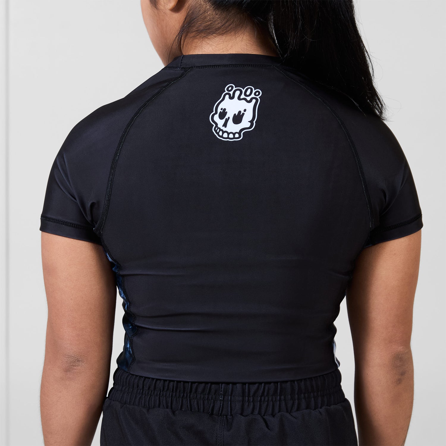 Back view of the Women’s Ash BJJ Rash Guard showing black design with white melting skull Rubber Bones logo near neckline.