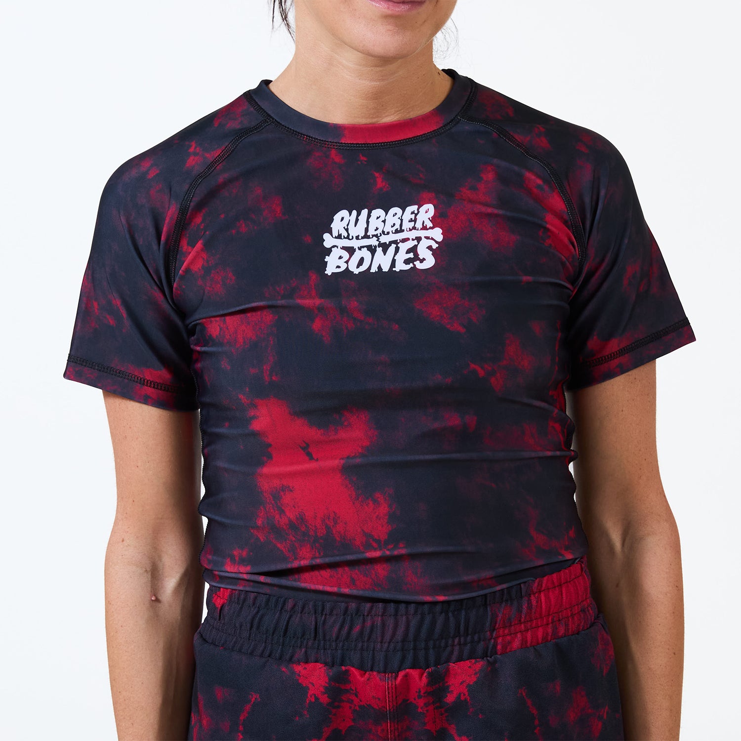 Model wearing the Women’s Ember BJJ Rash Guard featuring a red and black tie-dye pattern and white Rubber Bones logo.