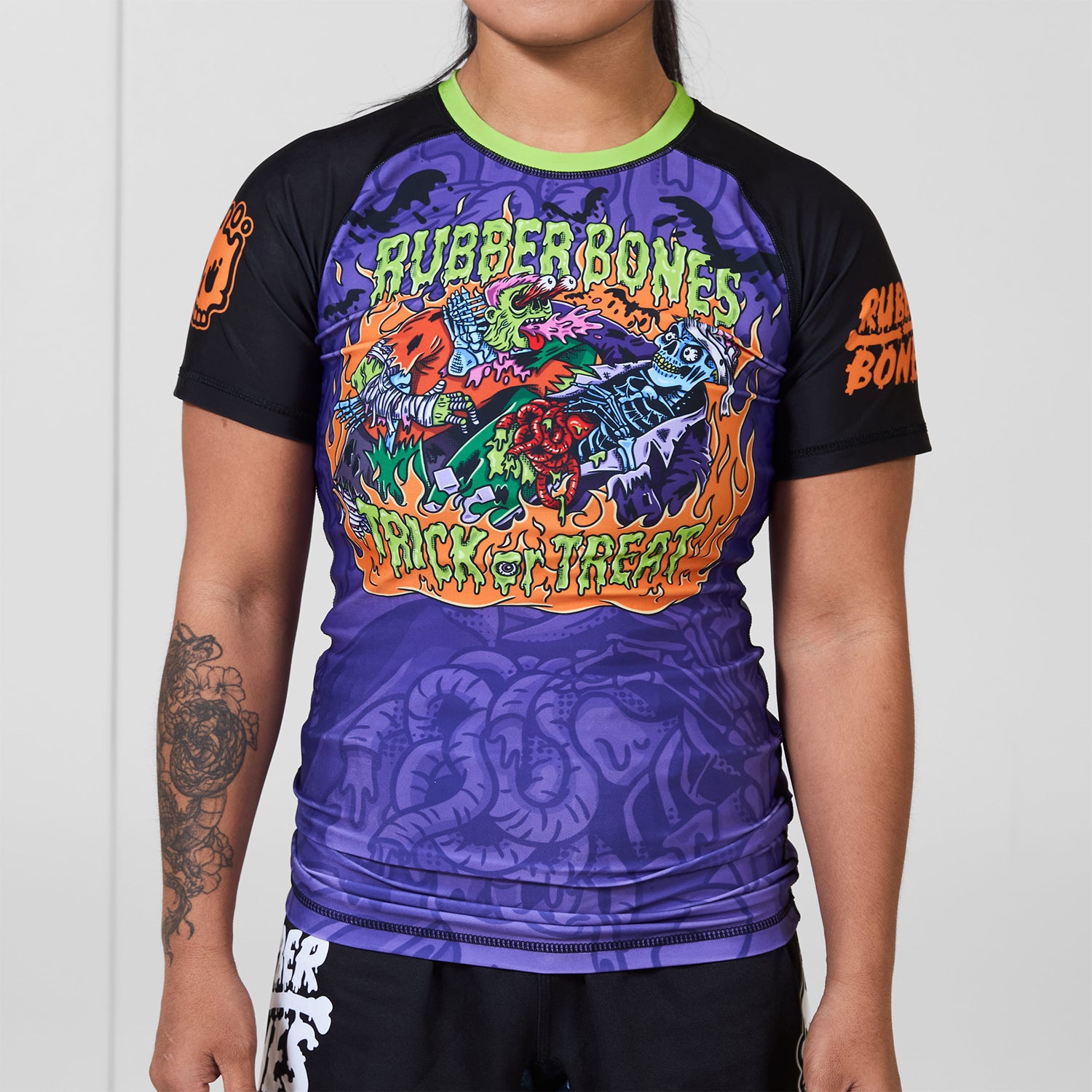 Front view of the Rubber Bones Trick or Treat women’s BJJ rash guard in purple, black, and neon green with bold monster artwork.