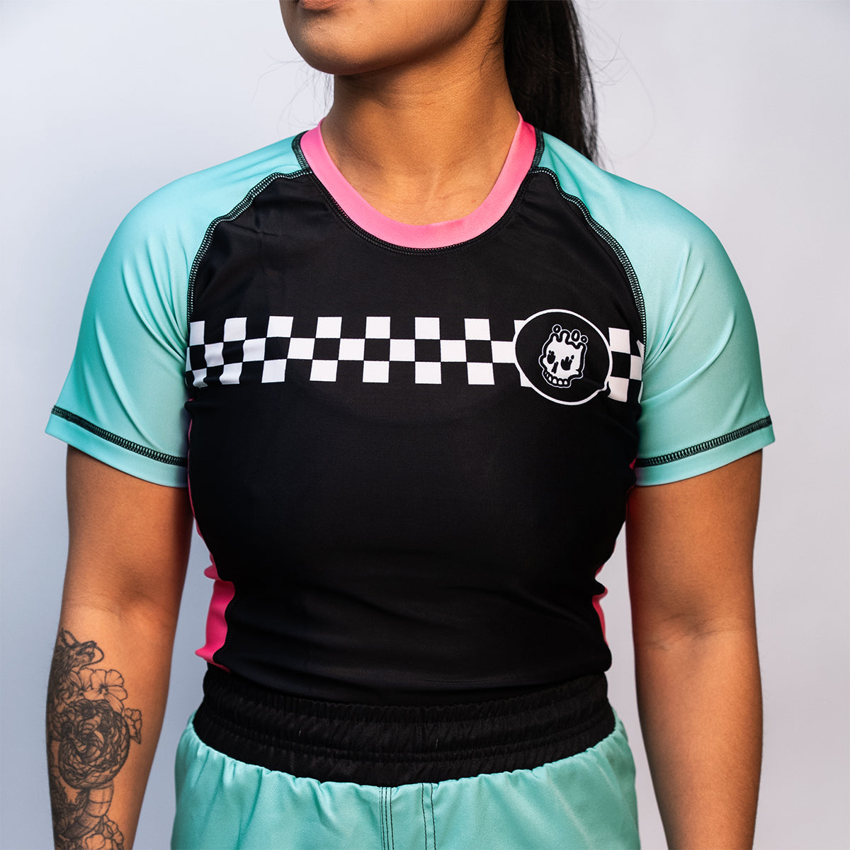 A woman wearing a short-sleeve rash guard with teal mint sleeves, black torso, and pink side panels. The front features a white checker stripe across the chest with the Rubber Bones skull logo on the left side.