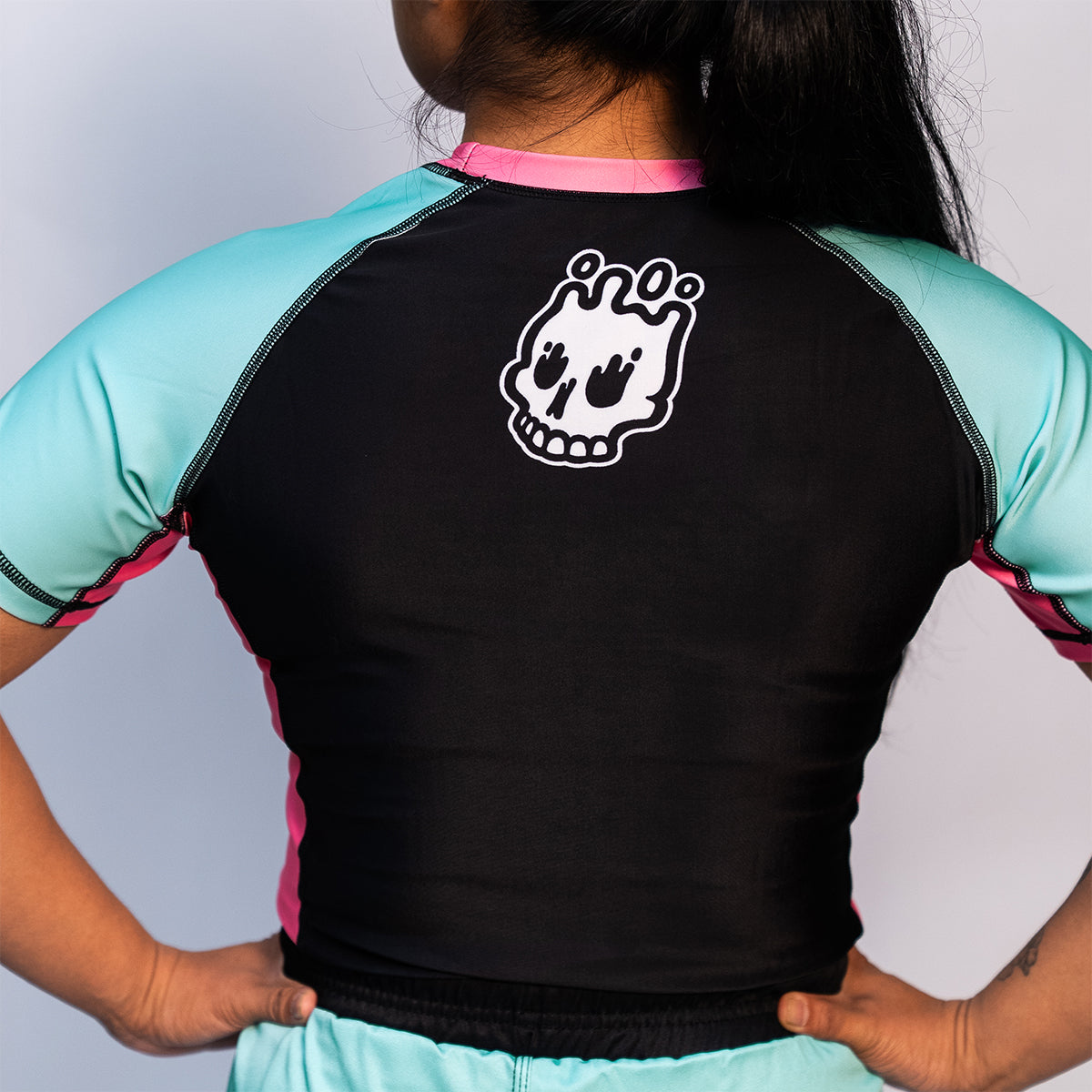Back view of the rash guard, showing a black back panel, teal mint sleeves, and pink side panels. The Rubber Bones white skull logo is printed at the upper center back.