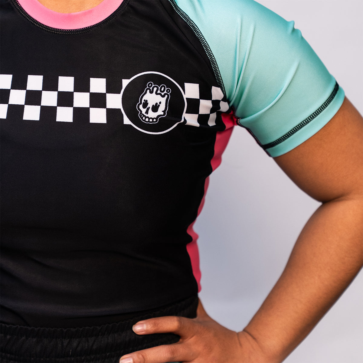 Close-up of the chest area showing the checkerboard stripe and Rubber Bones skull logo on the black torso of the rash guard. Teal sleeves and pink collar are also visible.