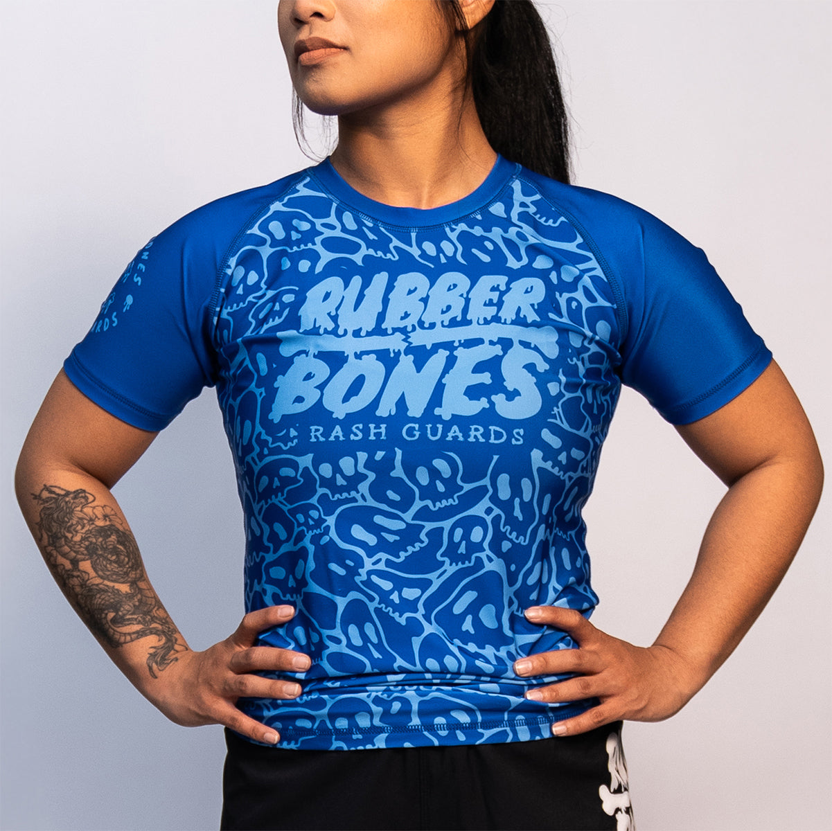 Woman wearing a blue ranked Brazilian Jiu-Jitsu rash guard with an all-over skull pattern. Features "Rubber Bones Rash Guards" logo in the center. Short sleeves with reinforced stitching.