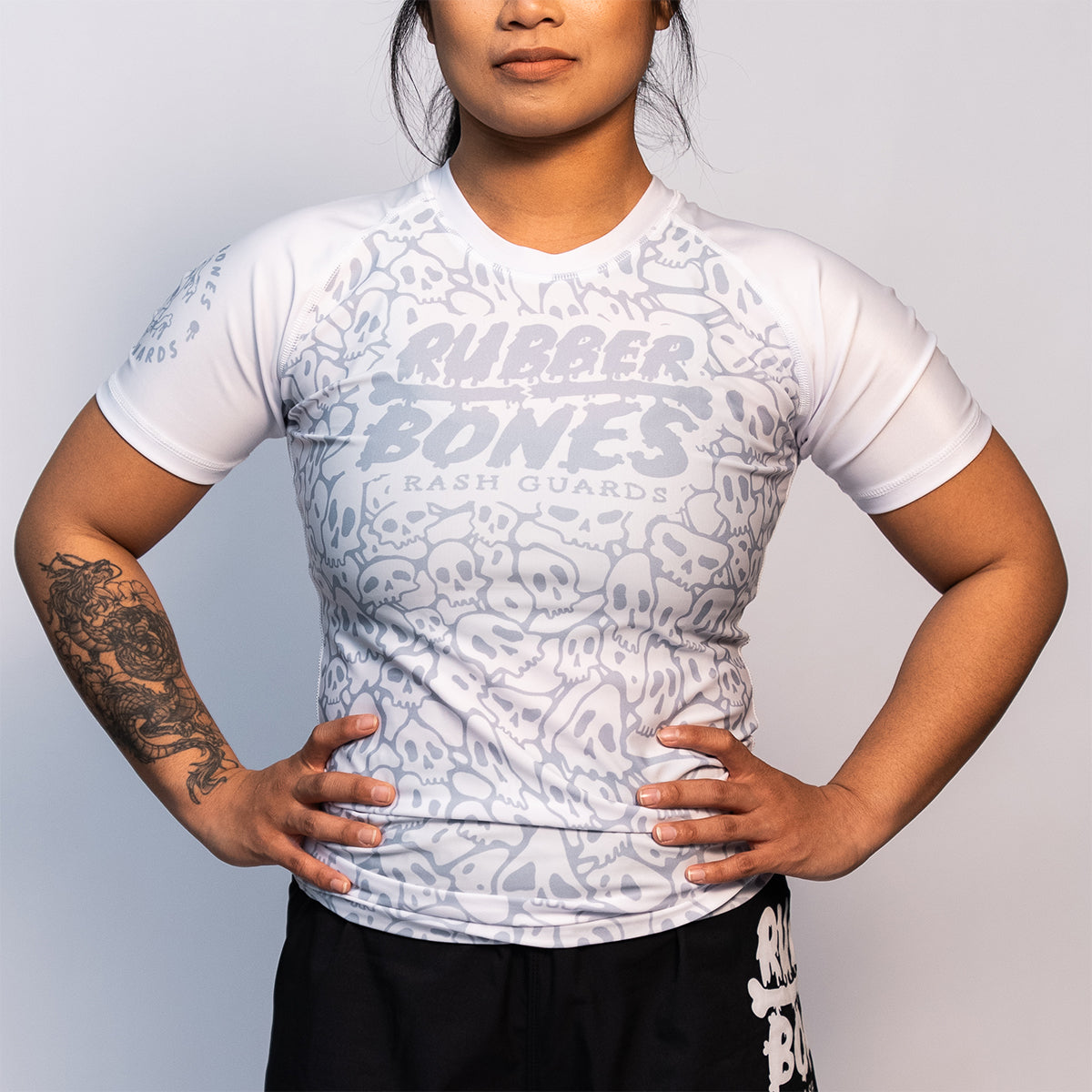 Front view of a women's white BJJ ranked rash guard featuring an all-over light gray skull pattern, short sleeves, and a fitted cut. The chest displays the "Rubber Bones Rash Guards" logo in gray.