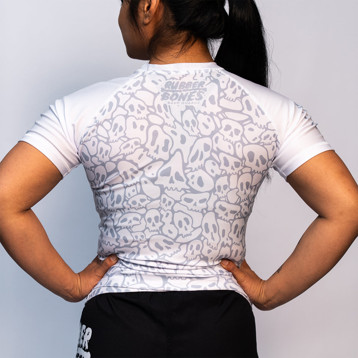 Back view of a women's white BJJ ranked rash guard with a light gray repeating skull pattern and a high neckline. The upper back features the "Rubber Bones Rash Guards" logo.