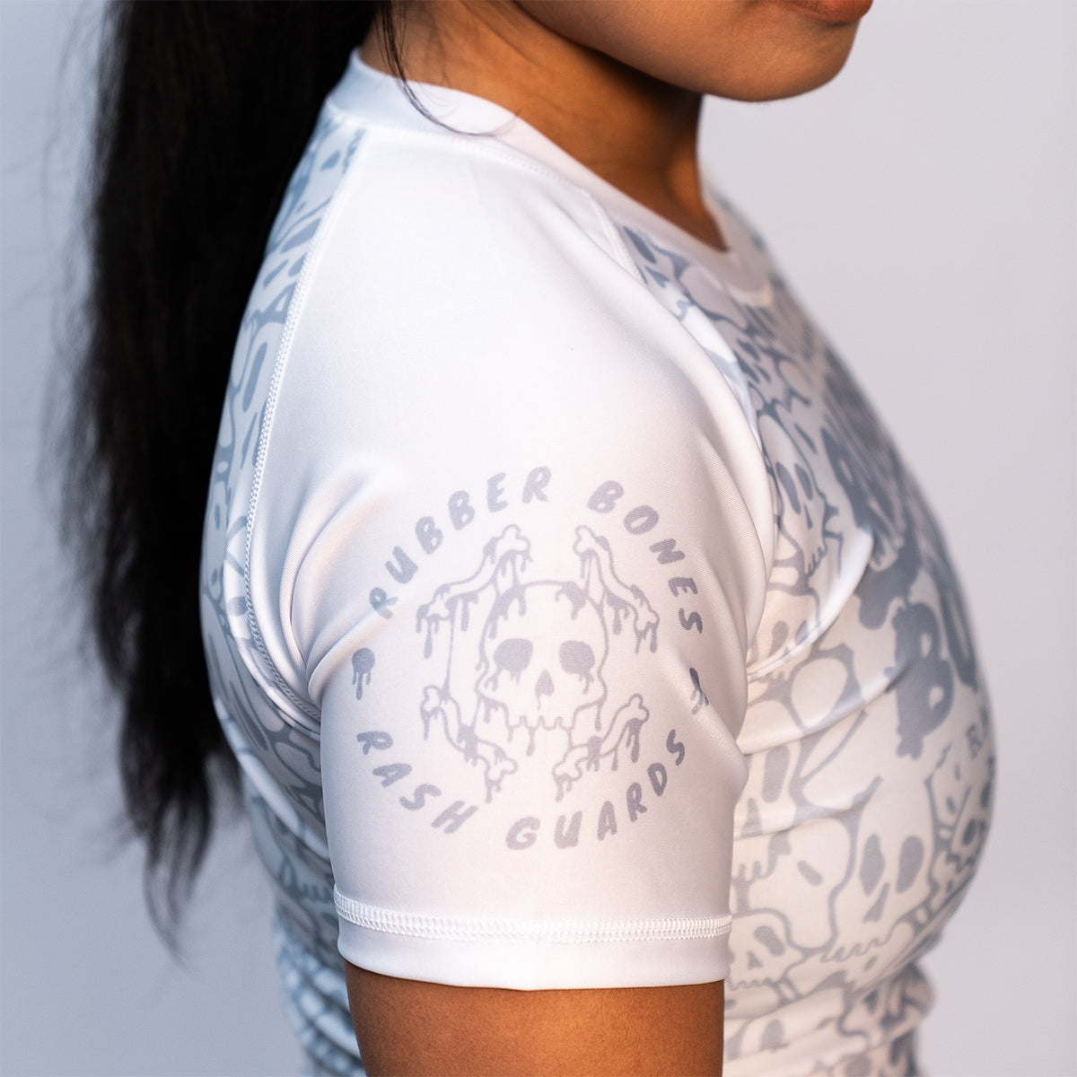 Side view of a women's white BJJ ranked rash guard showcasing the light gray skull pattern, short sleeves, and a dripping skull logo on the upper arm with "Rubber Bones Rash Guards" text encircling it.