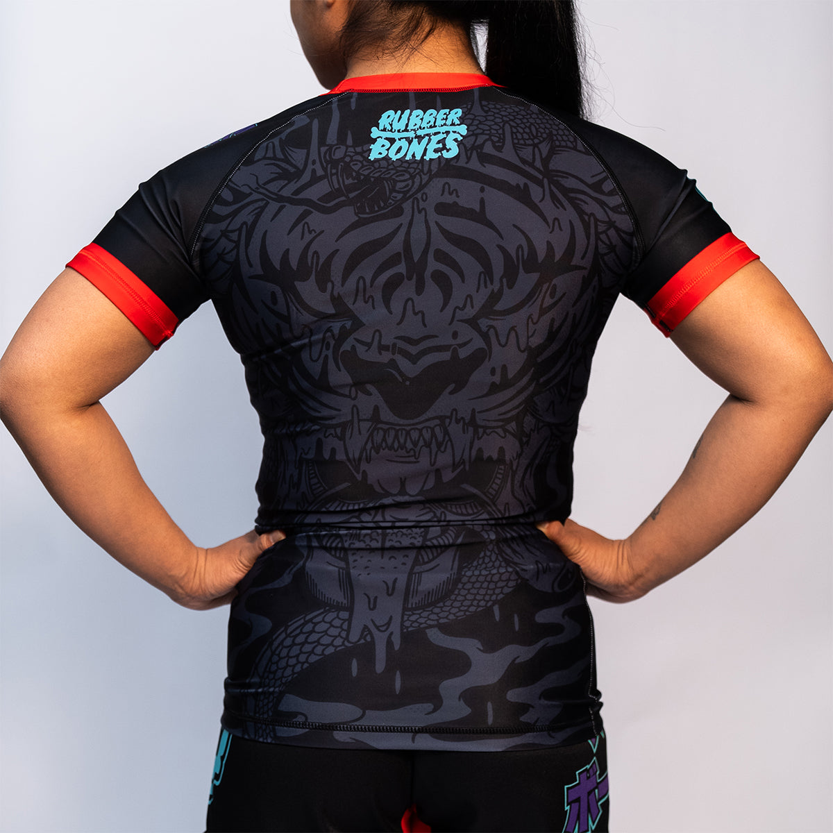 Back view of the Rubber Bones Tora Oni women's rash guard – Black no-gi rash guard featuring a tonal tiger-demon design, red sleeve cuffs, and Rubber Bones branding on the upper back.